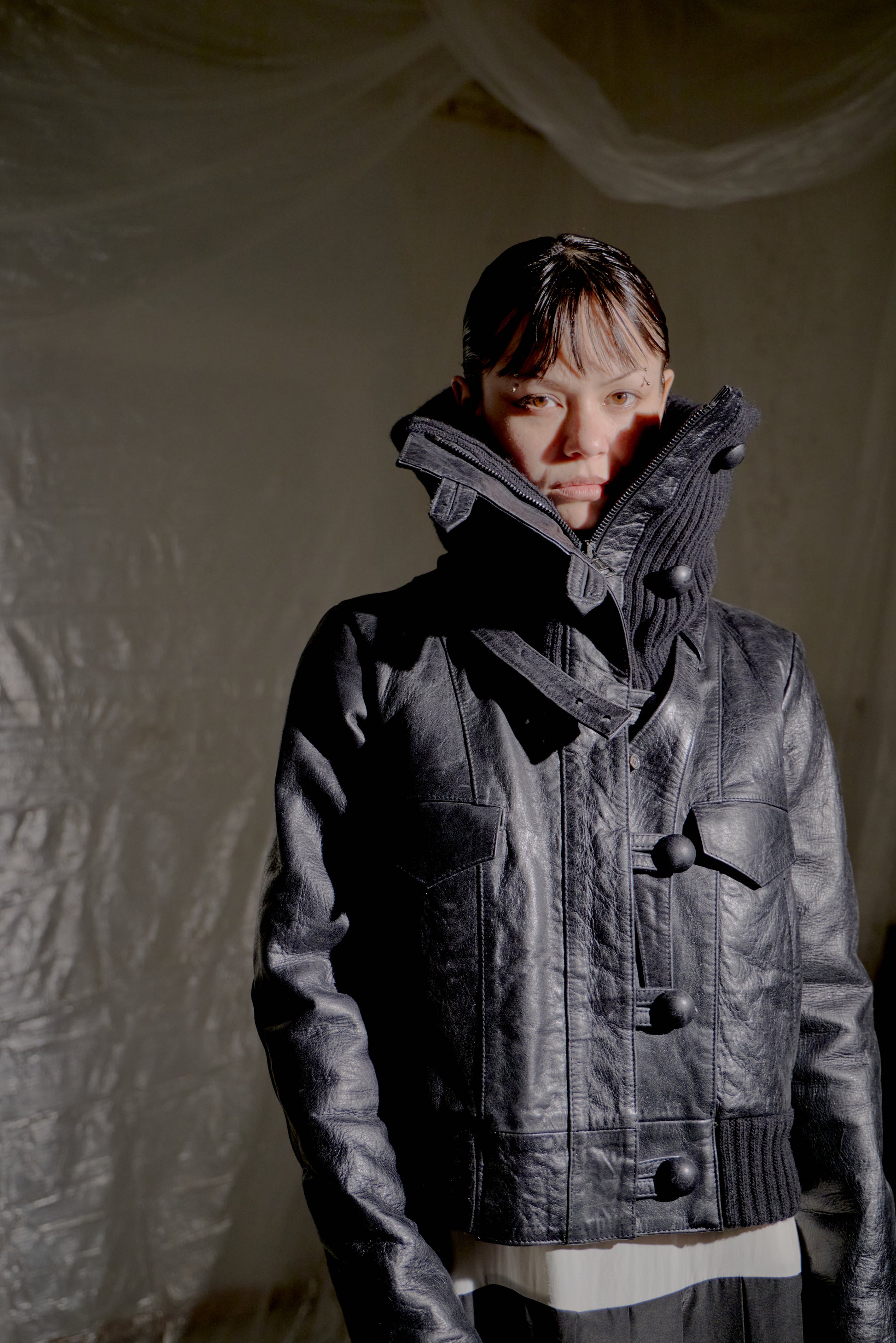 Person wearing a black Balenciaga leather jacket with a hood against a textured white wall.