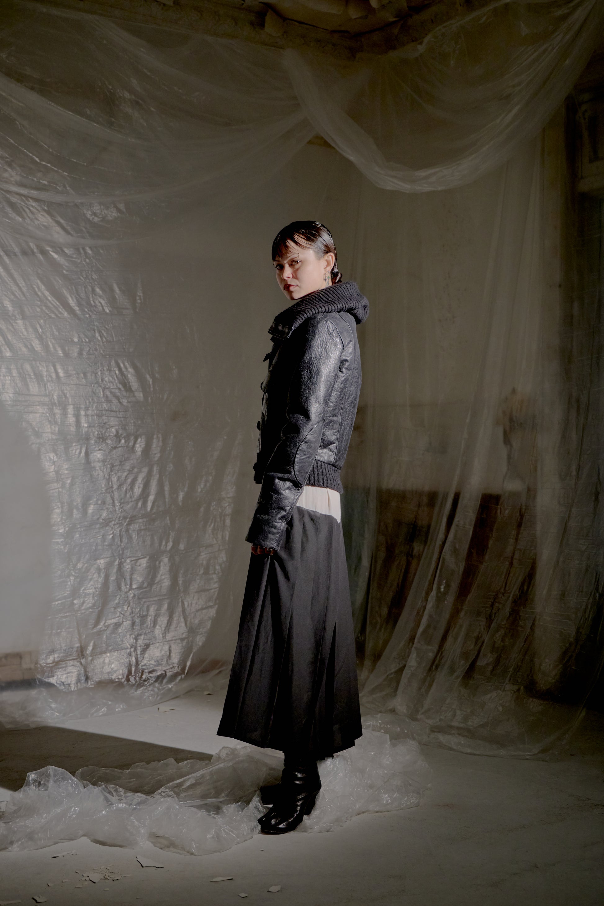 Person wearing a black leather Balenciaga jacket and skirt standing in a draped fabric-covered room.