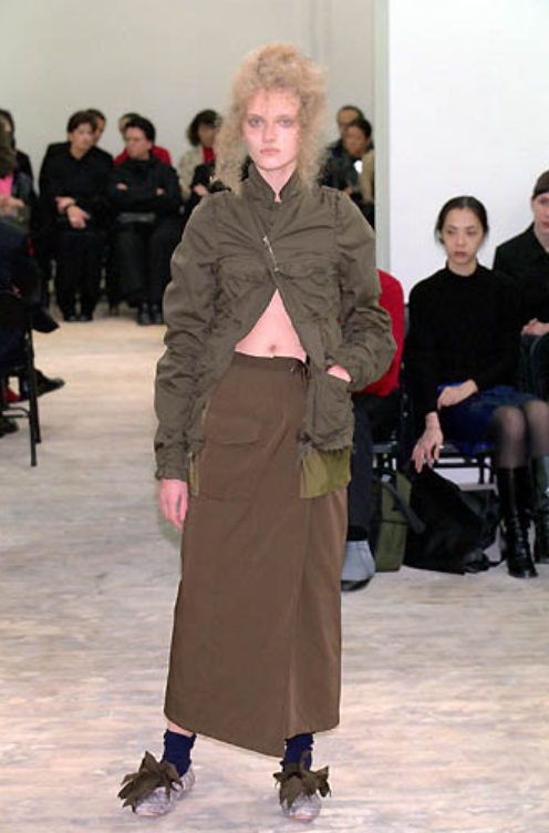 Model walks runway in an olive green top and brown skirt from Comme Des Garçons with bow-tied shoes. Audience in background observes. 