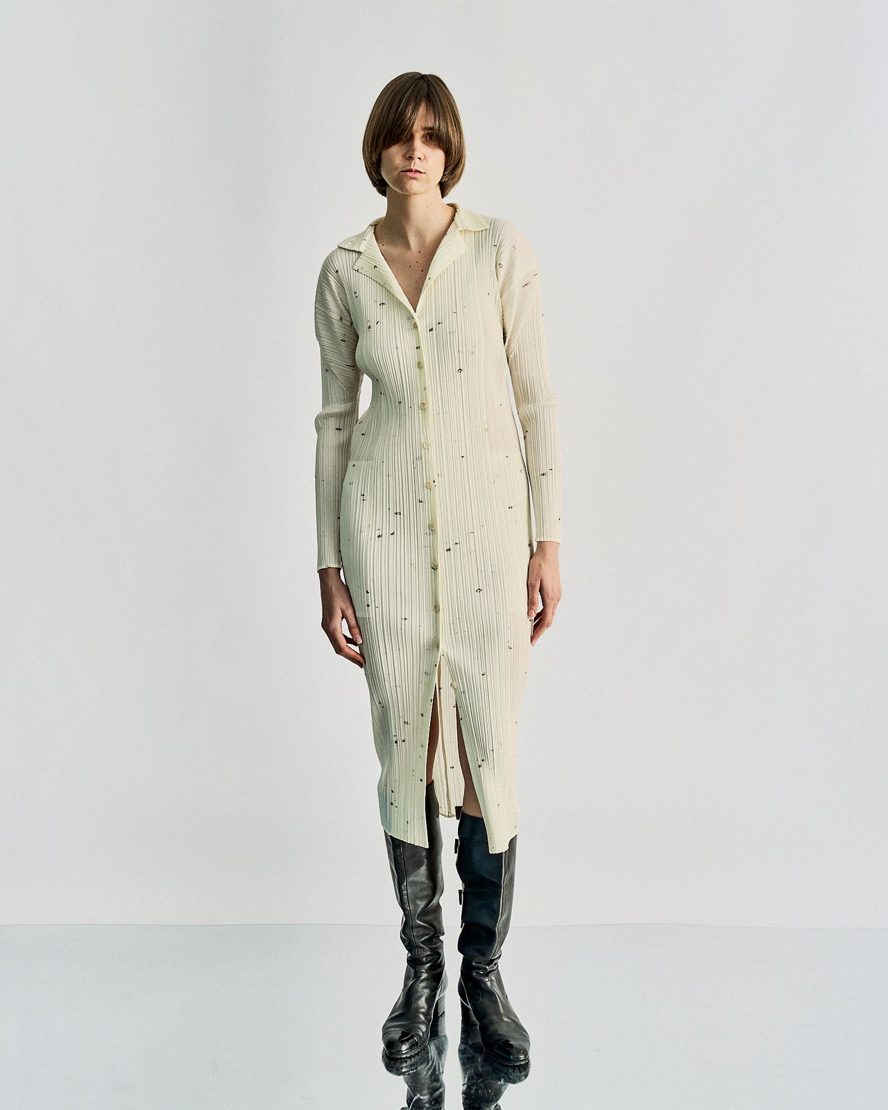 A person in a white, button-up Issey Miyake dress with delicate embellishments stands against a plain background. They wear tall black boots.