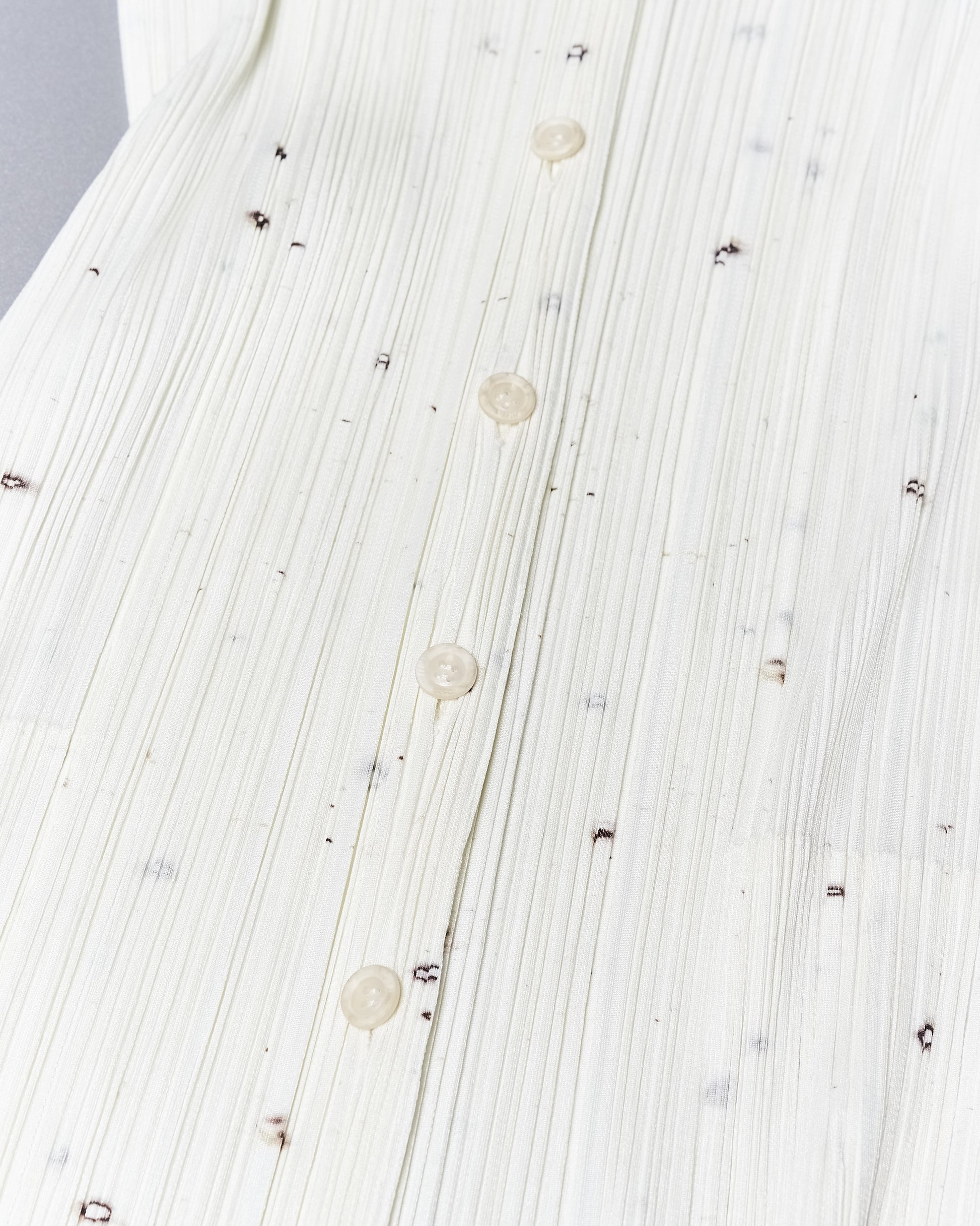 Close-up of a white, pleated fabric with subtle vertical lines and scattered small dark spots on an Issey Miyake dress. Transparent buttons are evenly spaced down the centre.