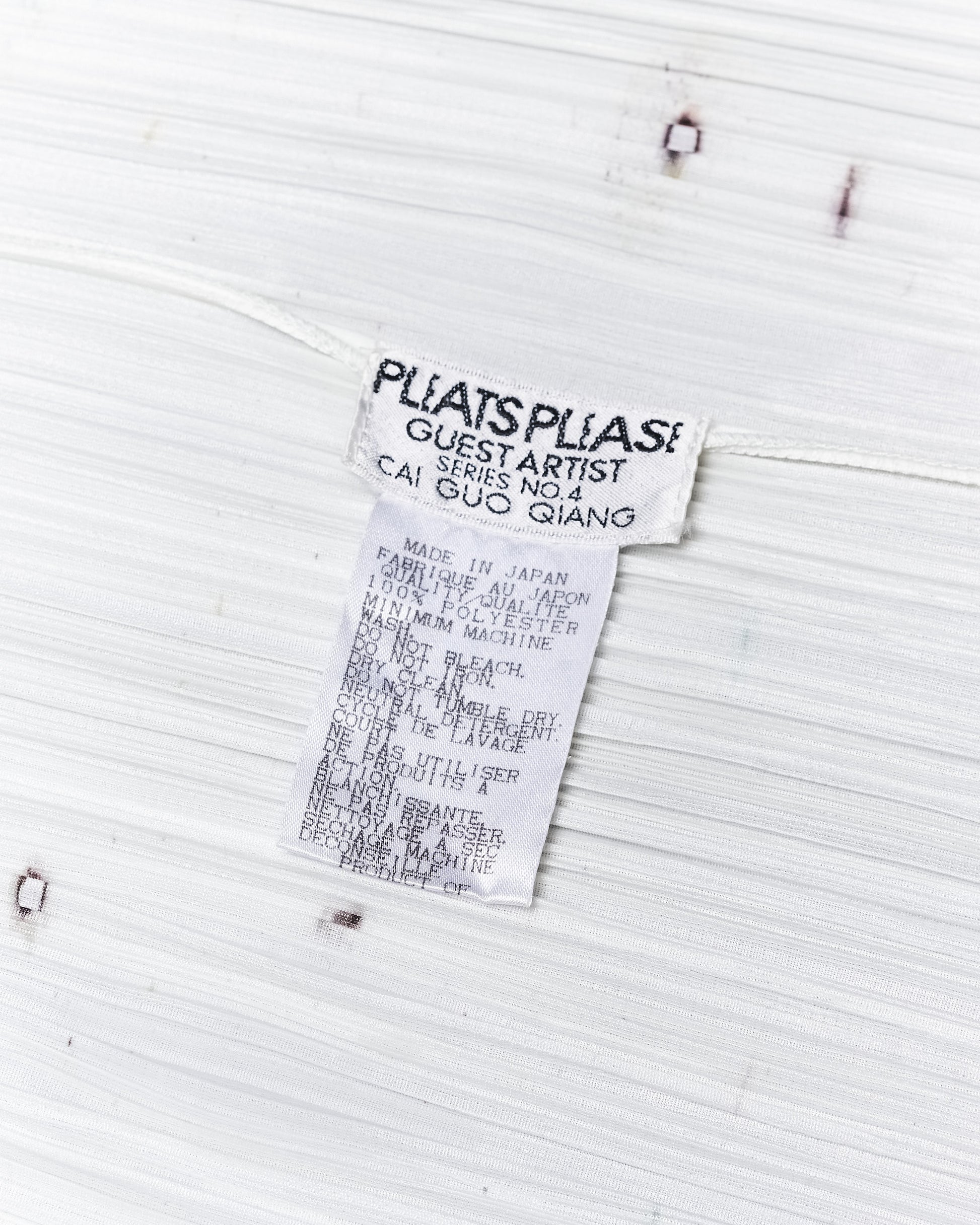 White fabric with a clothing label reads "Pleats Please, Guest Artist Series No.4, Cai Guo Qiang." Text details care instructions in both English and French.
