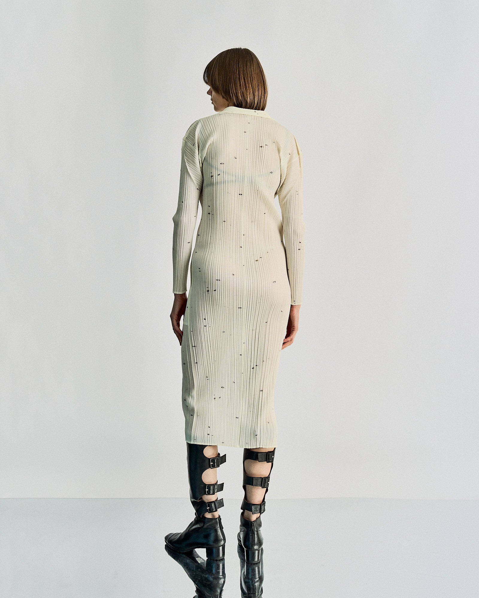 Aperson stands with their back to the camera in a minimalist setting, wearing a long, beige, button-up Issey Miyake dress with subtle patterns and tall black boots.