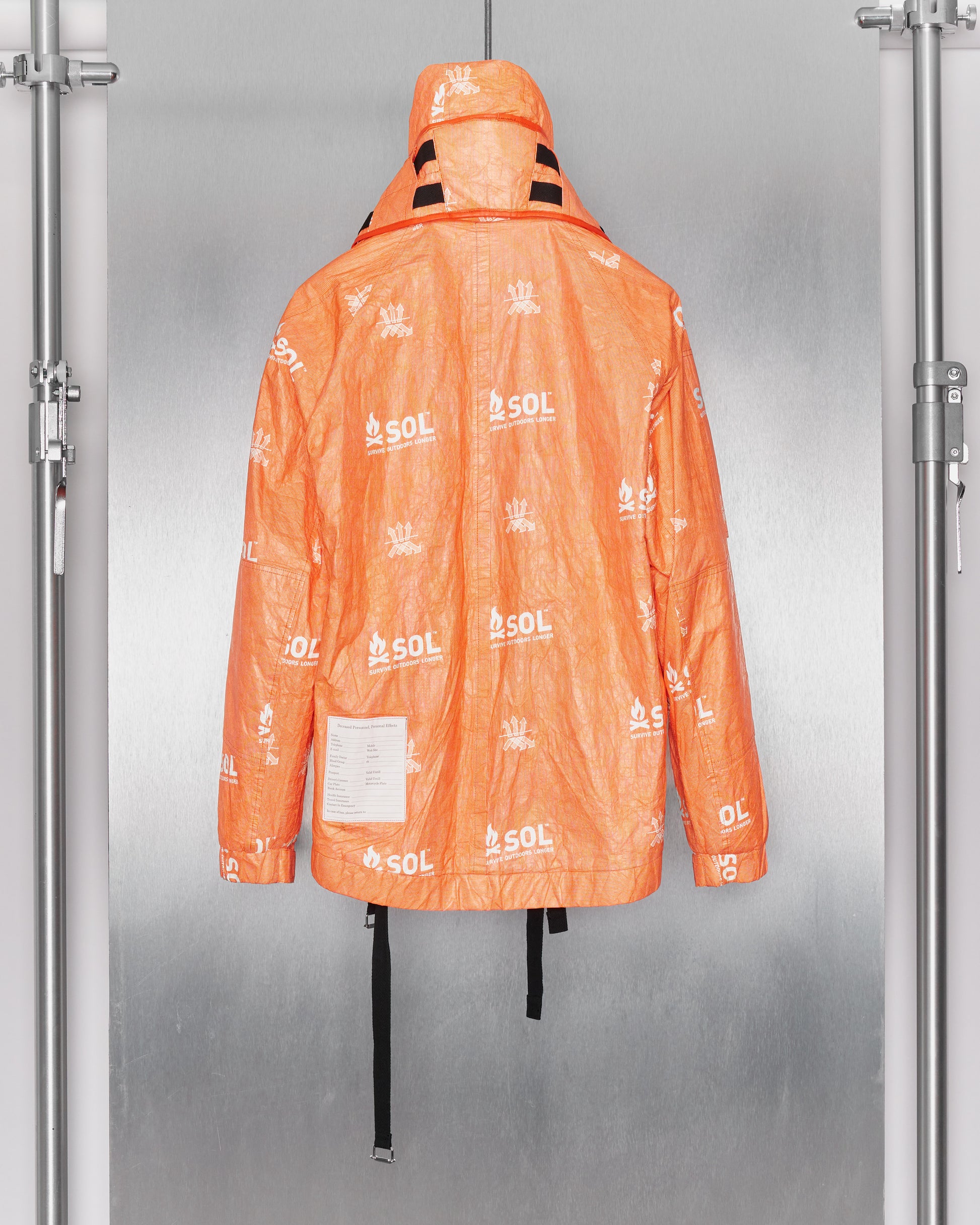 Back of orange jacket with hood up by TAKAHIROMIYASHITA TheSoloist covered in white logos, hanging on display against a metallic background. It has a hood, black straps, and a patch with writing.