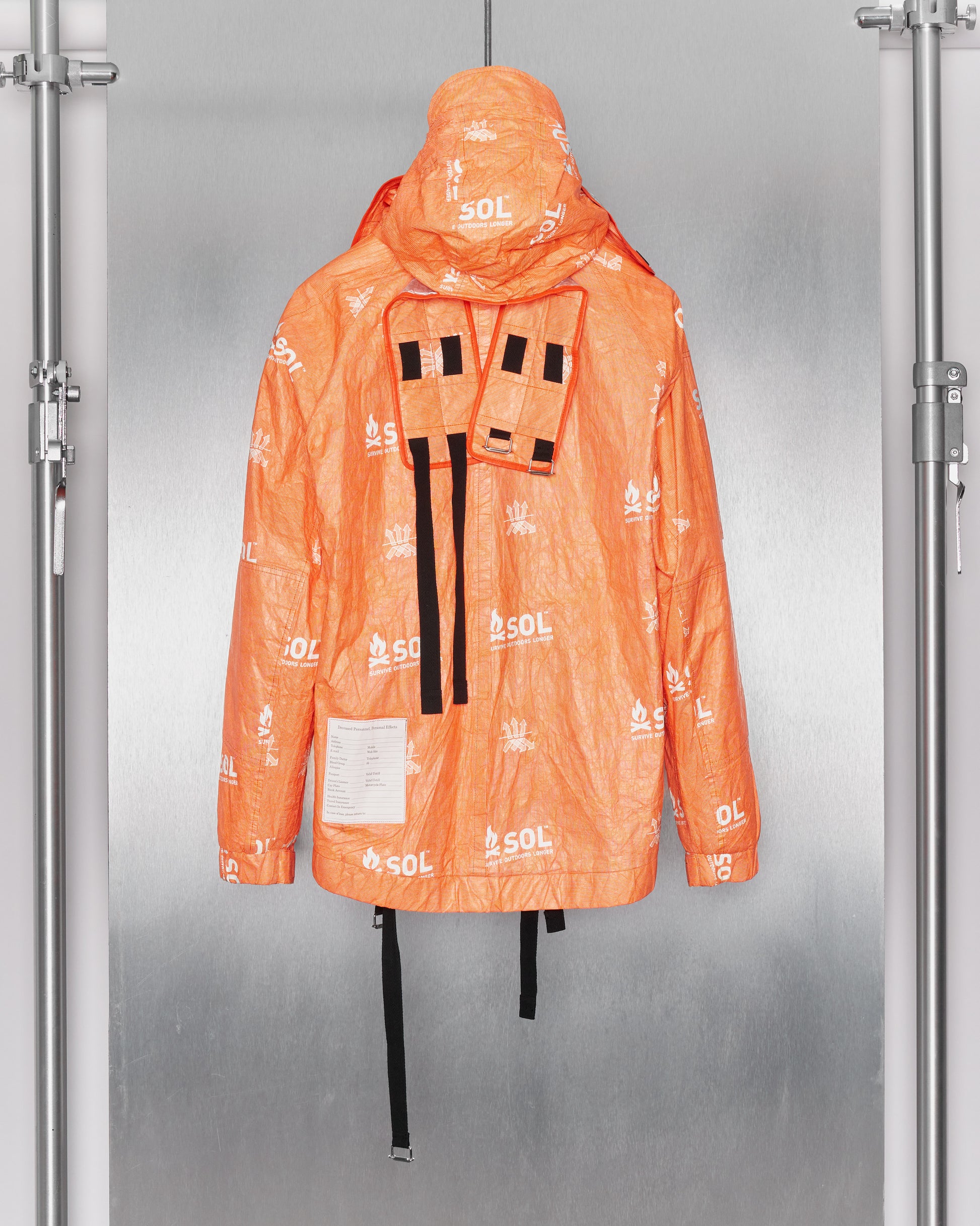 Back of orange jacket by TAKAHIROMIYASHITA TheSoloist covered in white logos, hanging on display against a metallic background. It has a hood, black straps, and a patch with writing.
