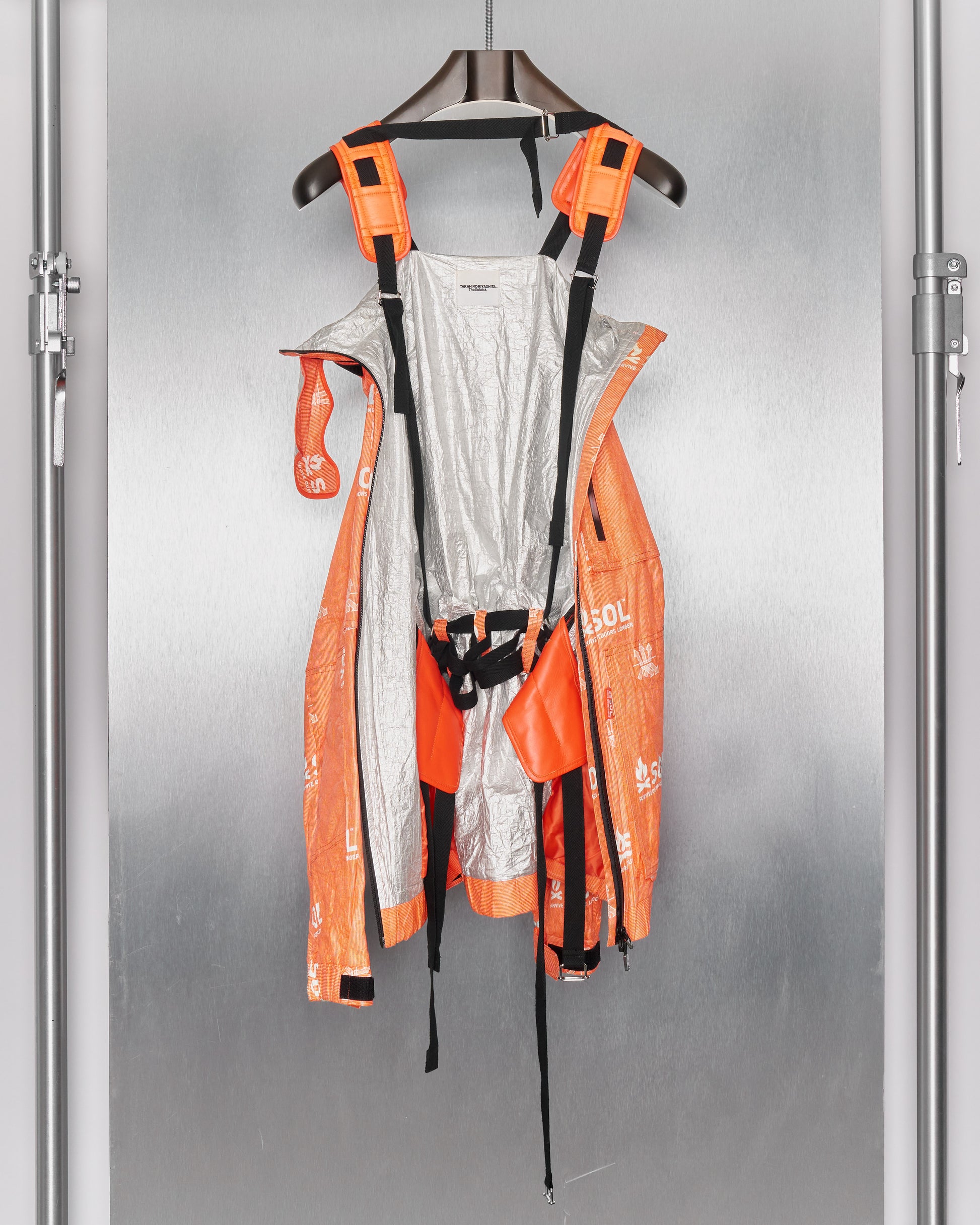 A futuristic, orange and grey harness jacket by TAKAHIROMIYASHITA TheSoloist hangs on a brown hanger in front of a metallic, industrial-style background.