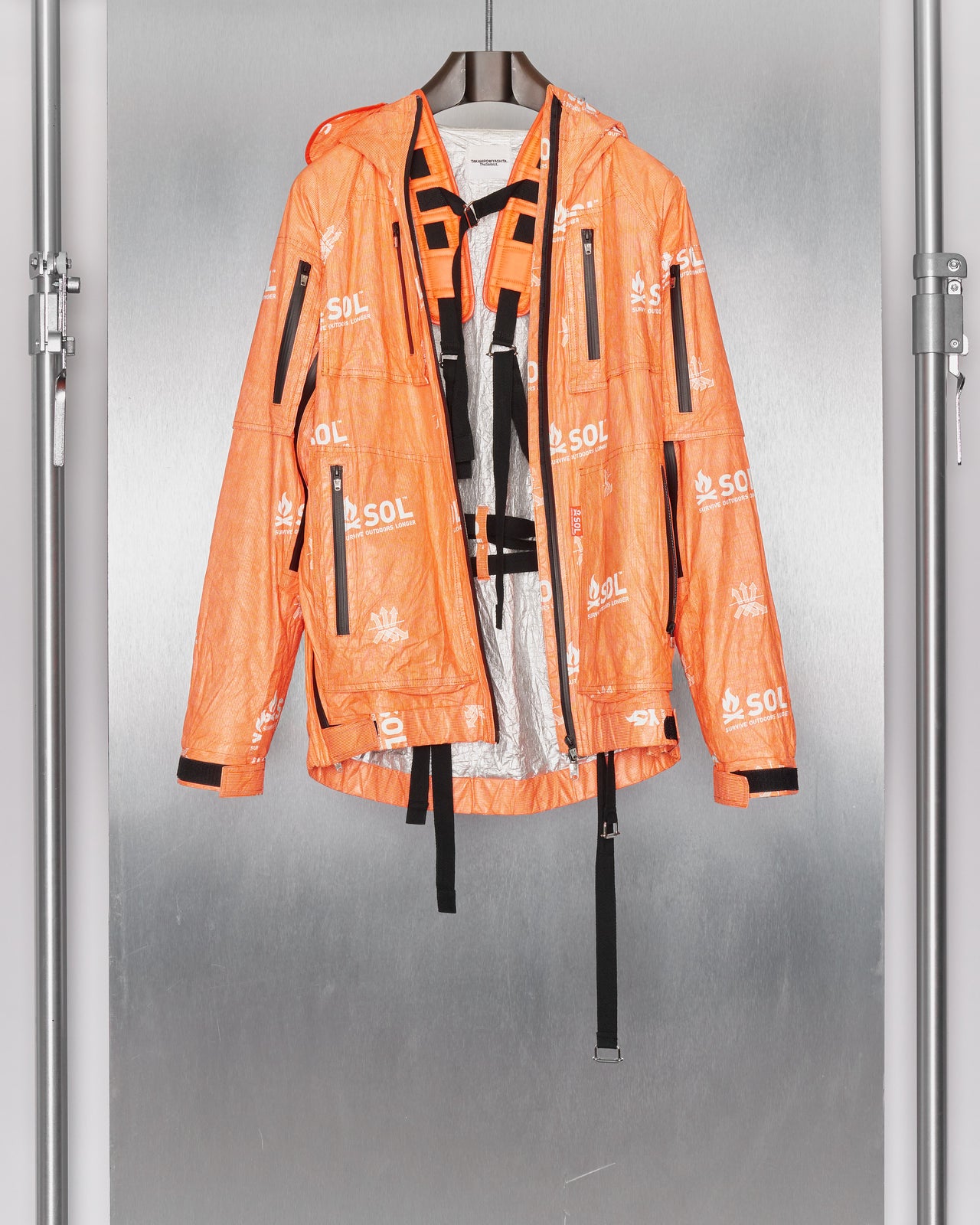 A model wearing an orange hooded jacket by TAKAHIROMIYASHITA TheSoloist and matching face cover walks down a dimly lit runway, exuding a mysterious, futuristic ambiance.