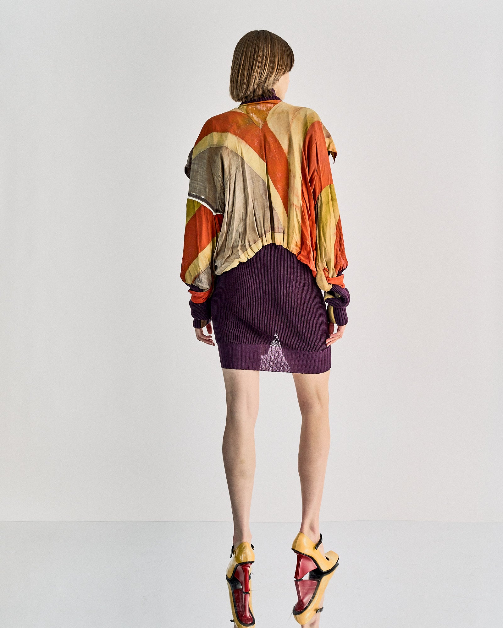 A person stands facing away, wearing a colourful Vivienne Westwood bomber jacket and a purple skirt. They are on high heels. 