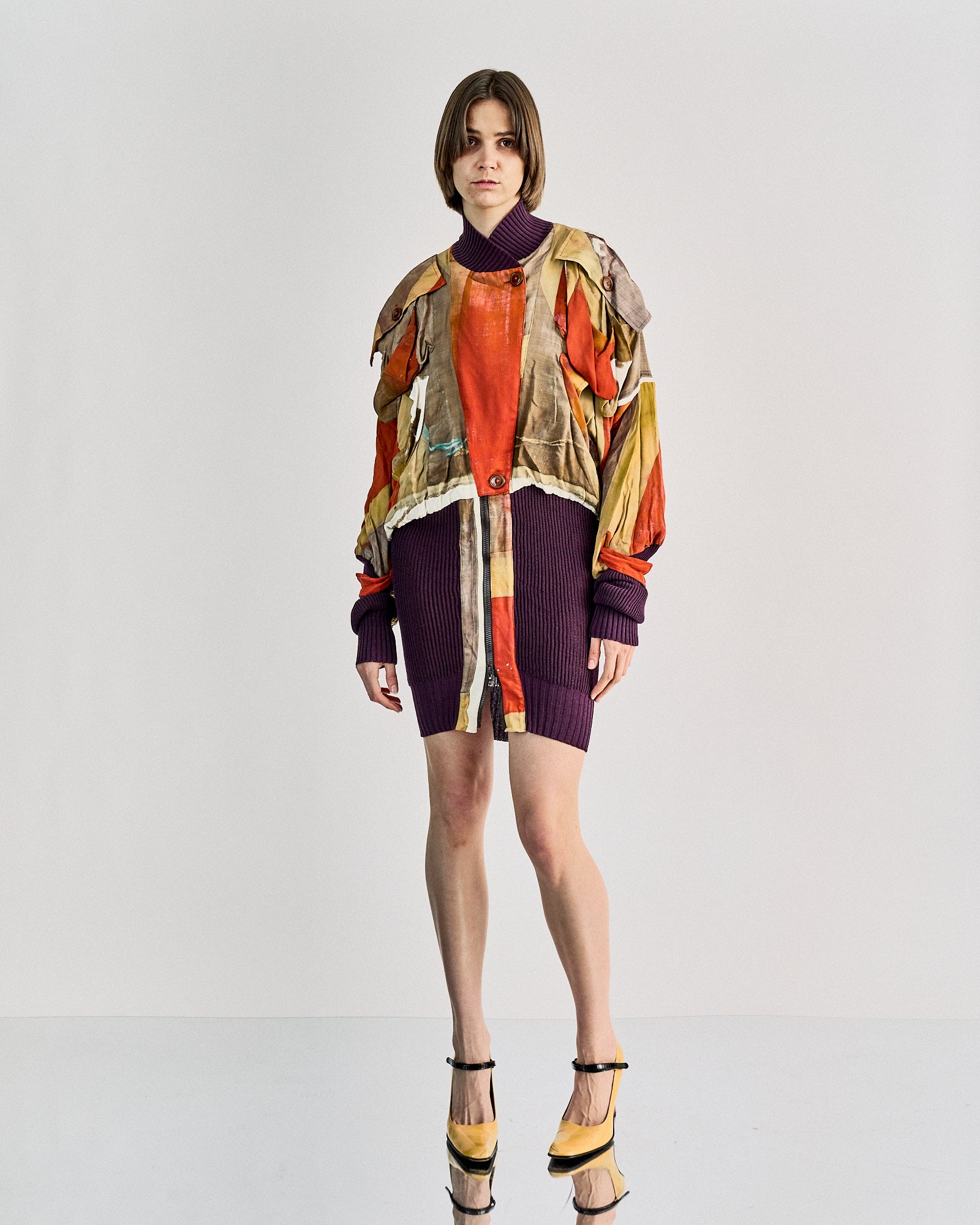 A woman stands in a studio wearing a colourful, oversized Vivienne Westwood jacket in earth tones over a deep purple knit dress. 